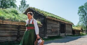 Kvinne i folkedrakt foran en tradisjonell torvtakshytte