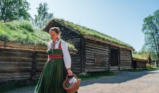 Kvinne i folkedrakt foran en tradisjonell torvtakshytte